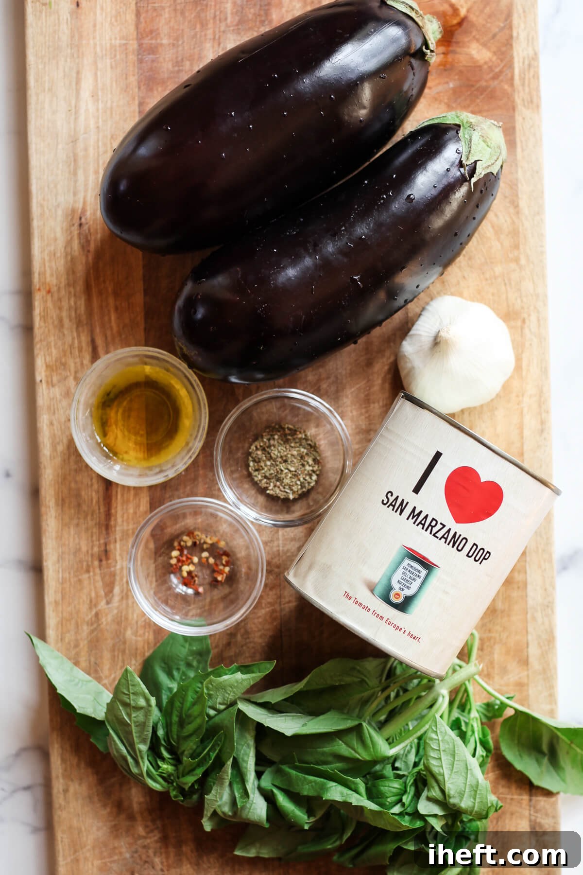 Two eggplants, a can of san marzano dop tomatoes, olive oil, garlic, and herbs on a wood cutting board. 