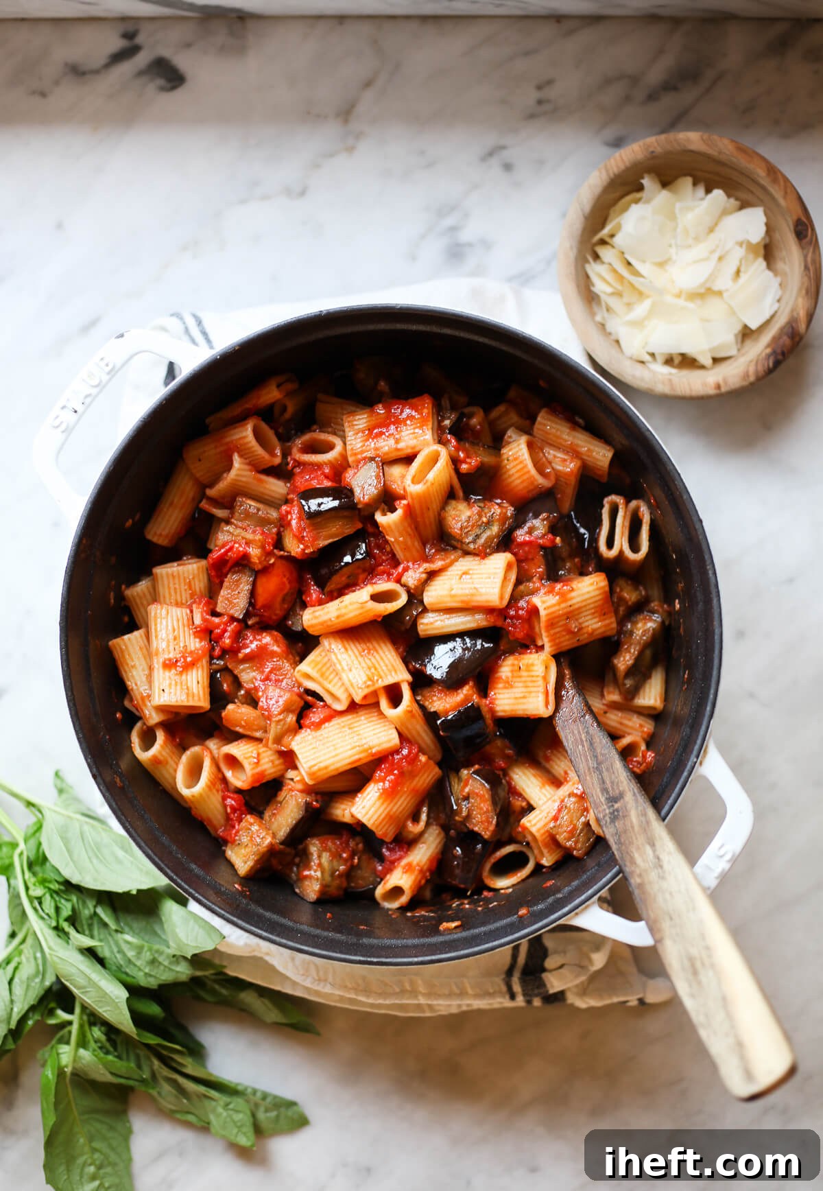A white Staub pot filled with Sicilian pasta alla norma recipe with eggplant ragu and tomatoes. The pot sits on a marble countertop with fresh basil and Parmesan cheese on the side. 