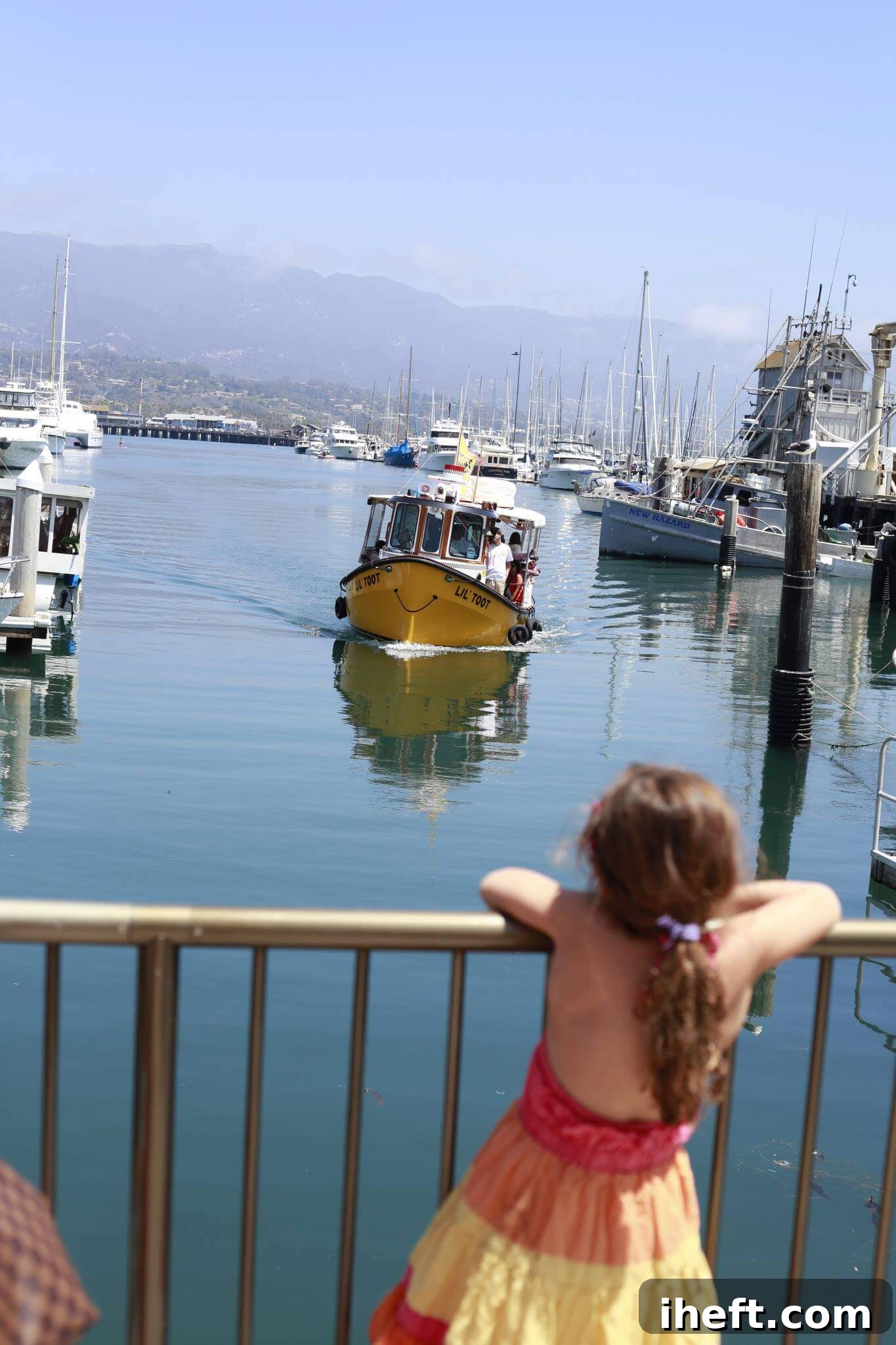A cute yellow boat in the Santa Barbara harbor approaches a little girl wearing a bright orange, pink, and yellow dress. 