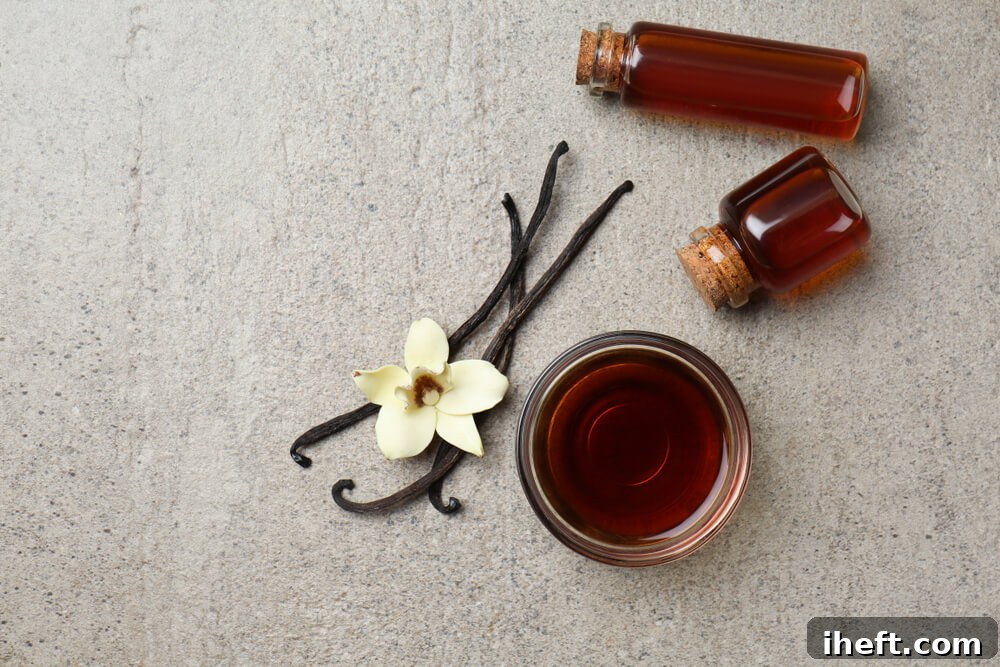 Three vanilla beans on a concrete countertop with a vanilla flower and small glasses of vanilla extract. 