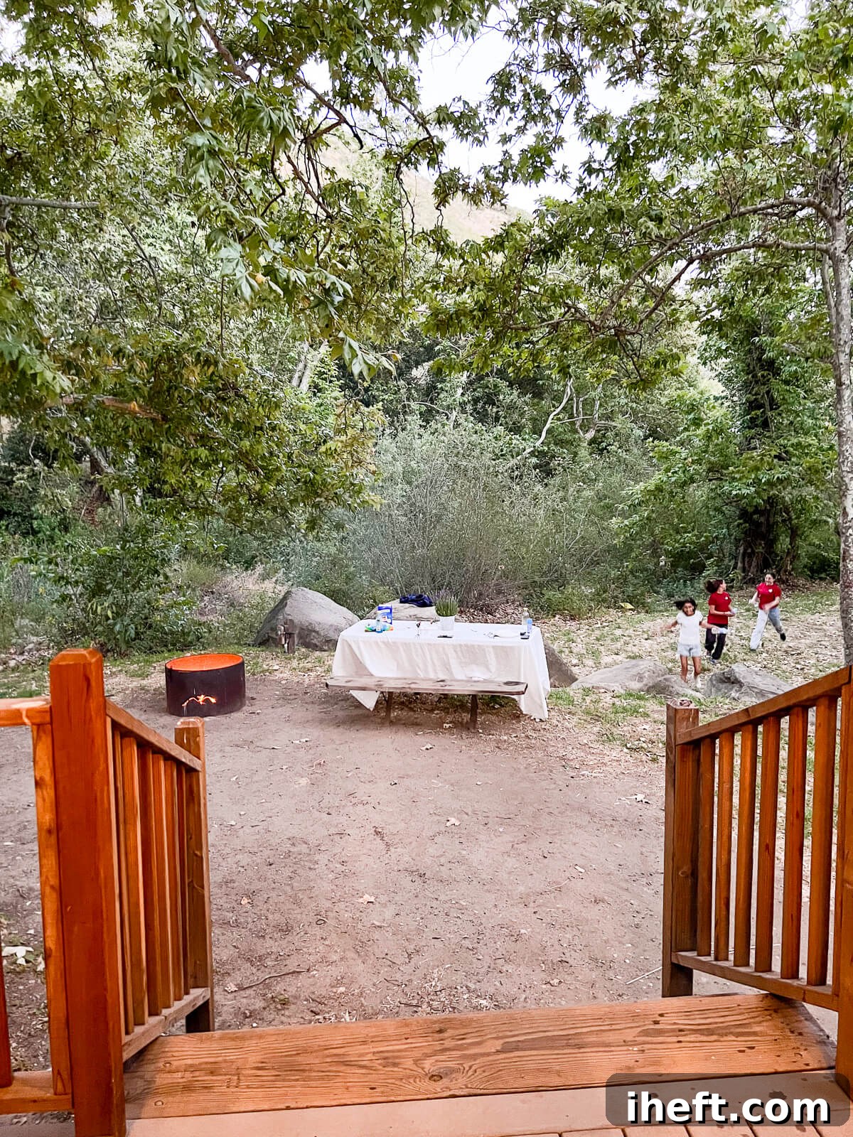 Three cheerful girls joyfully running and playing outside a cabin at the El Capitan Canyon resort. A rustic picnic table is invitingly set near a warmly glowing fire pit, perfect for evening gatherings.