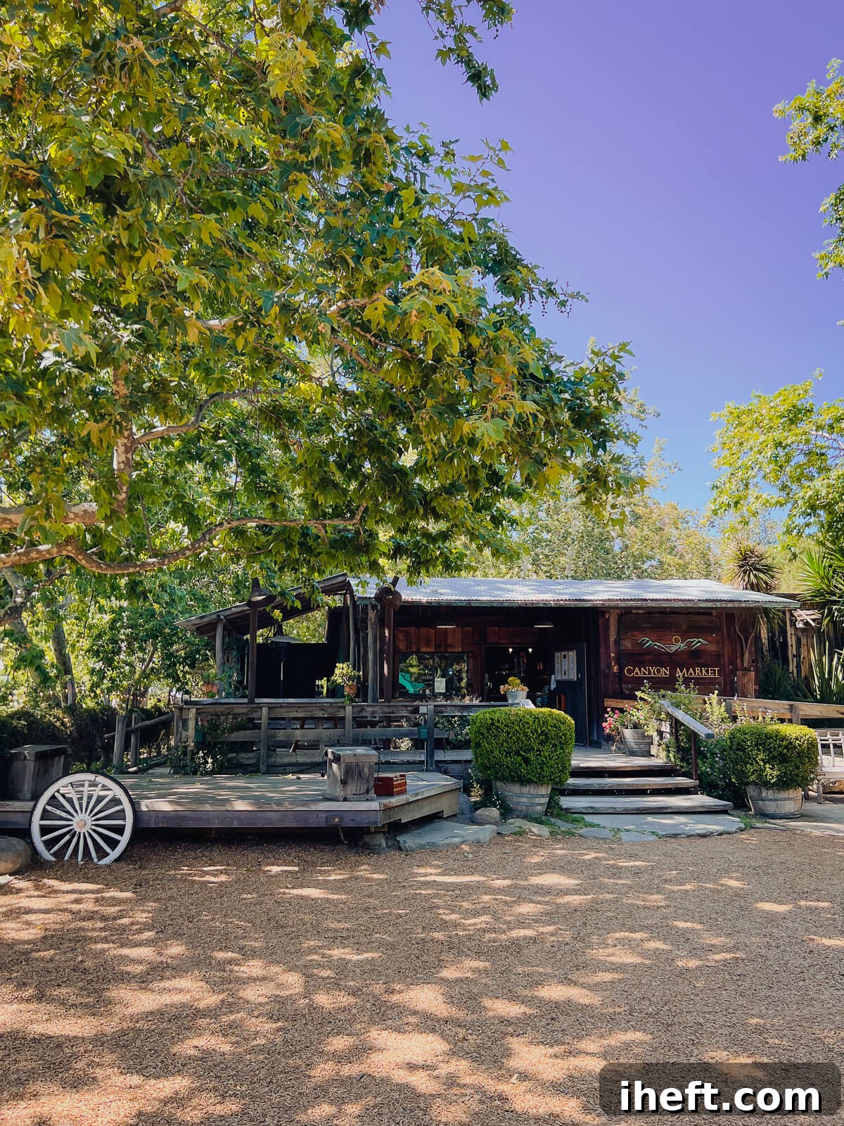 A quaint cedar general store, labeled "Canyon Market," shaded by majestic oak trees. A rustic wagon wheel rests beside a wooden stage, hinting at community events and gatherings.
