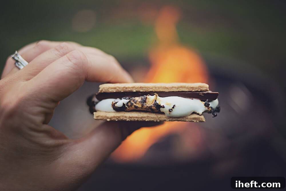 A close-up shot of a perfectly crafted s'more, featuring golden-brown roasted marshmallows and rich chocolate sandwiched between crunchy graham crackers, held in a hand in front of a warm campfire. 