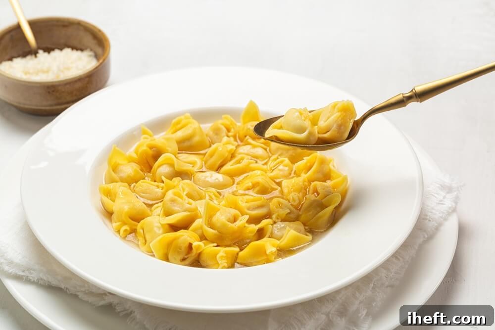 Tortellini in brodo (tortellini in broth) served in a white bowl, with a spoon gently lifting two tortellini, and a small bowl of grated cheese in the background.