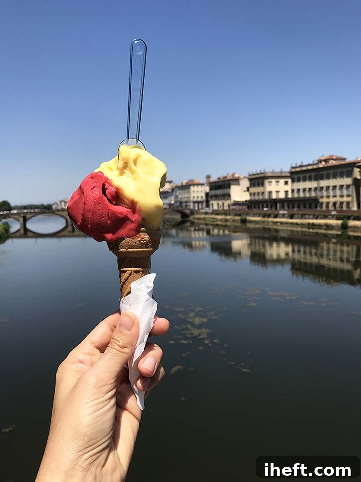 A hand holds a delightful cone of passion fruit and raspberry gelato with a small spoon, set against the scenic backdrop of the Arno river in Florence.