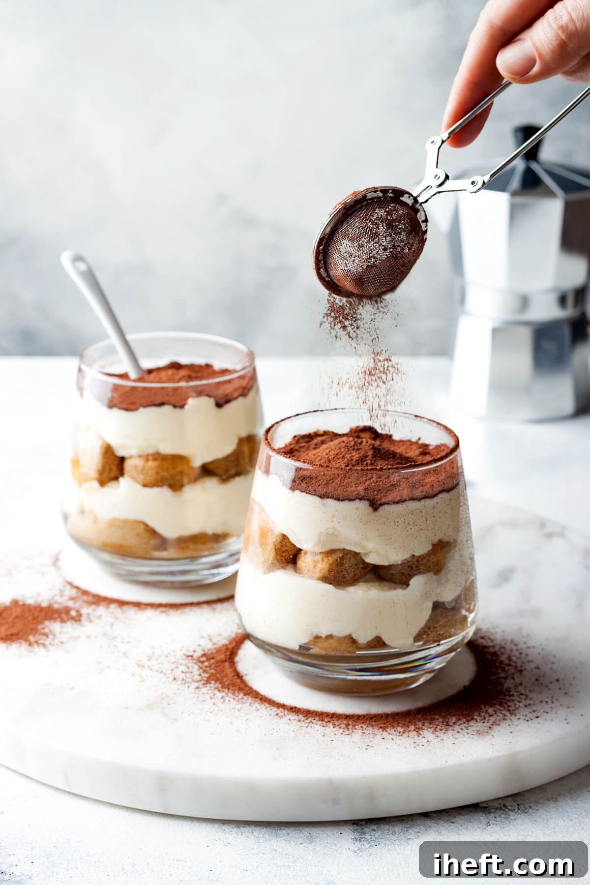 Cocoa powder being gently sifted over two individual glasses of homemade tiramisu, revealing layers of ladyfingers and creamy filling, with a moka pot in the background.