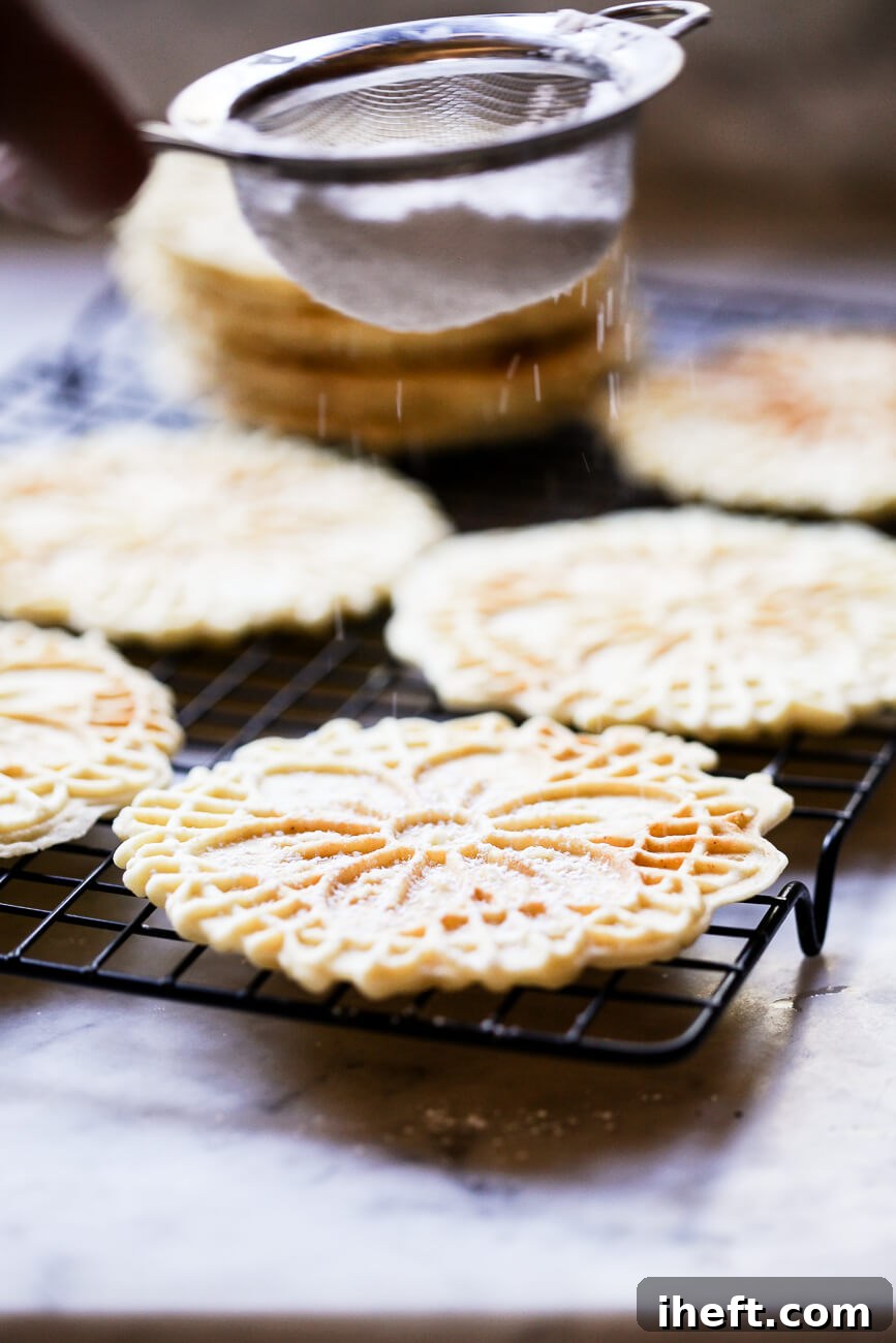 Powdered sugar being delicately sifted over homemade Pizzelle cookies, cooling on a rack, creating a beautiful snowy finish.