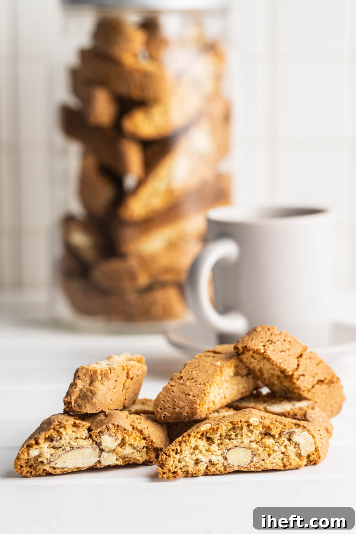 A charming pile of almond Cantucci biscotti resting on a white countertop, with a jar of more Cantucci and a coffee mug in the background, suggesting a delightful coffee break.