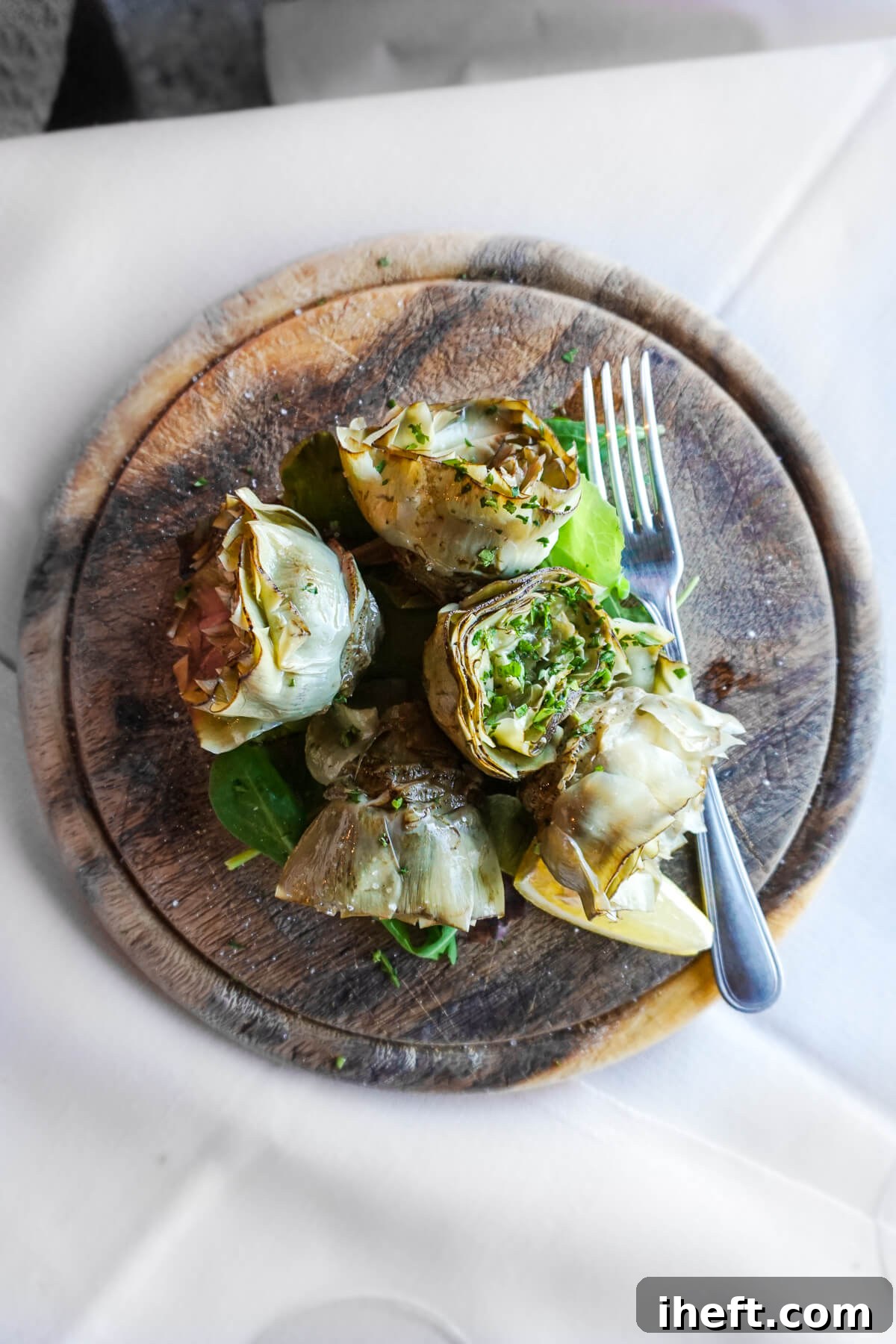 A round wooden board laden with Roman farmer-style Italian artichokes, garnished with fresh herbs, captured at a restaurant in Tuscany.
