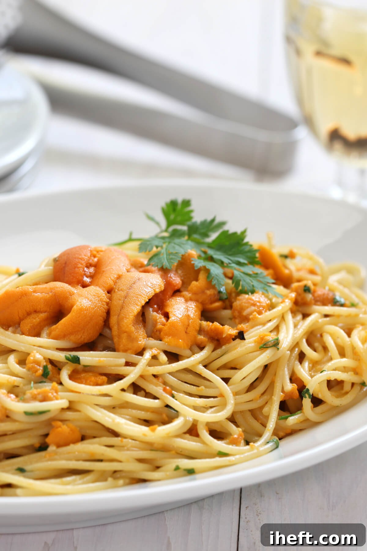 A white pasta bowl filled with creamy uni pasta (ricci di mare) topped with parsley. A glass of white wine in the background. 