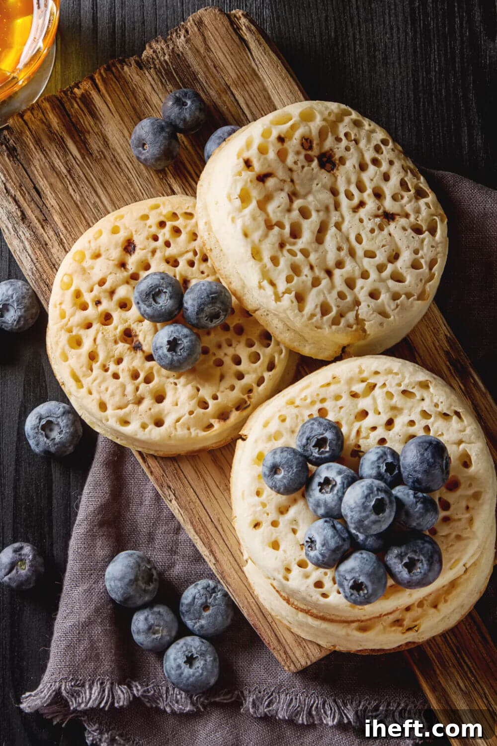 Three perfectly crafted English crumpets, beautifully garnished with fresh blueberries, resting on a rustic wooden cutting board.