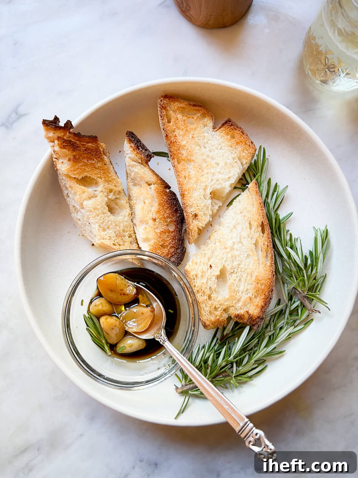 5 cloves of garlic confit in a small glass bowl of balsamic vinegar on a white plate with pieces of toast. 