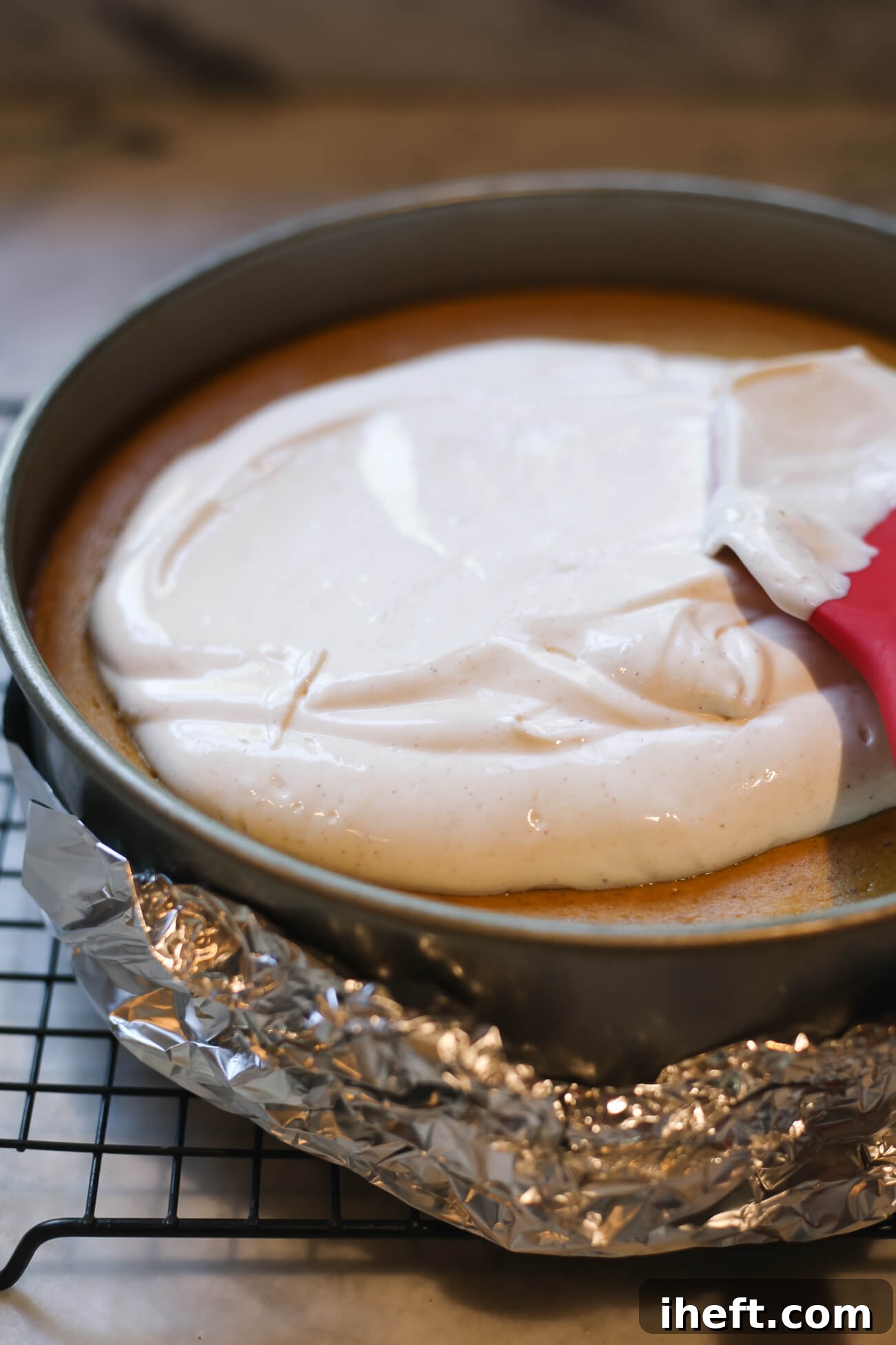 A vibrant red spatula expertly spreading a creamy, tangy sour cream topping over a freshly baked pumpkin cheesecake.