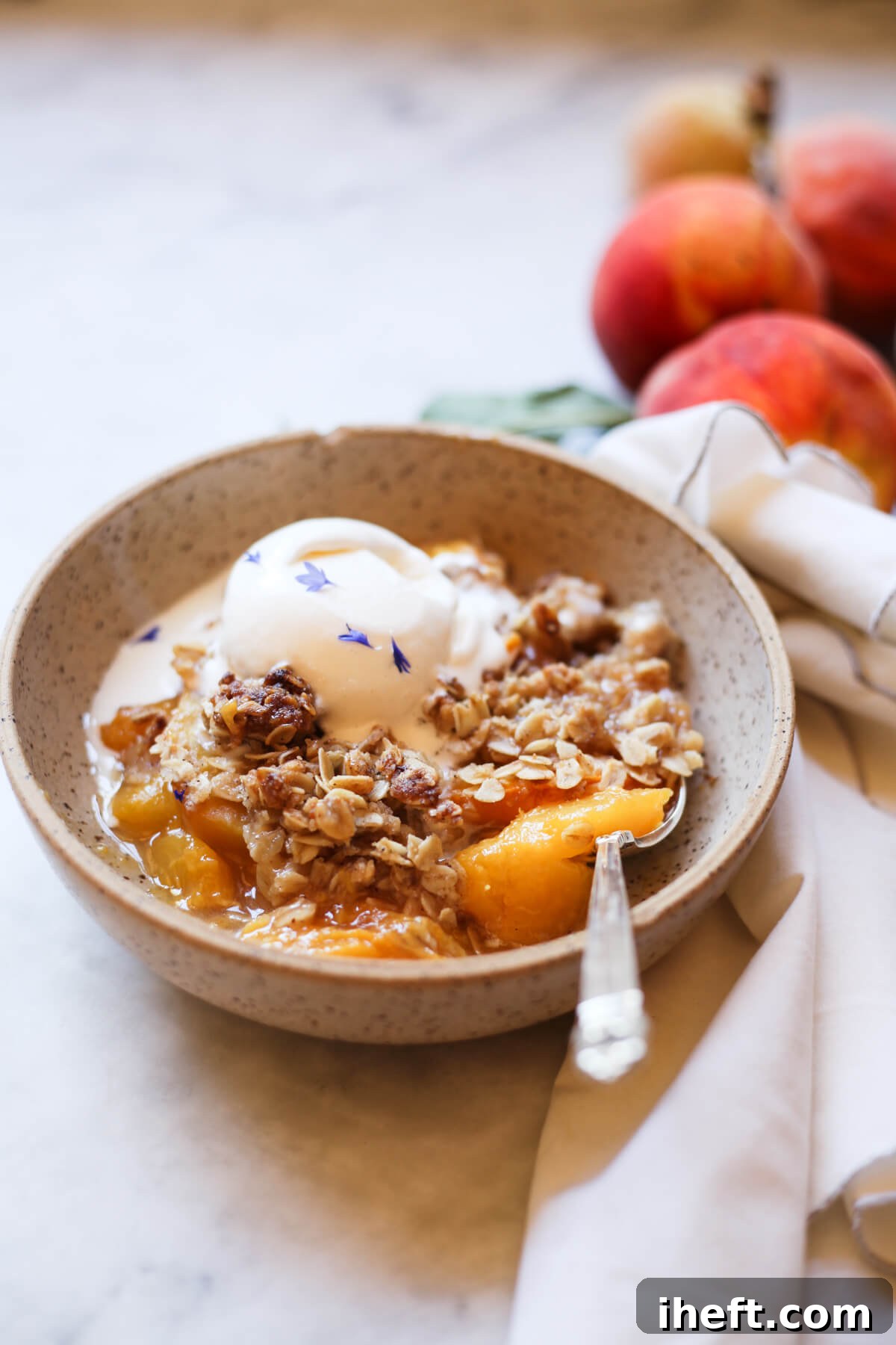 Peach crisp with oat crumble topping and a scoop of vanilla ice cream in an earthenware bowl on a white marble countertop. 