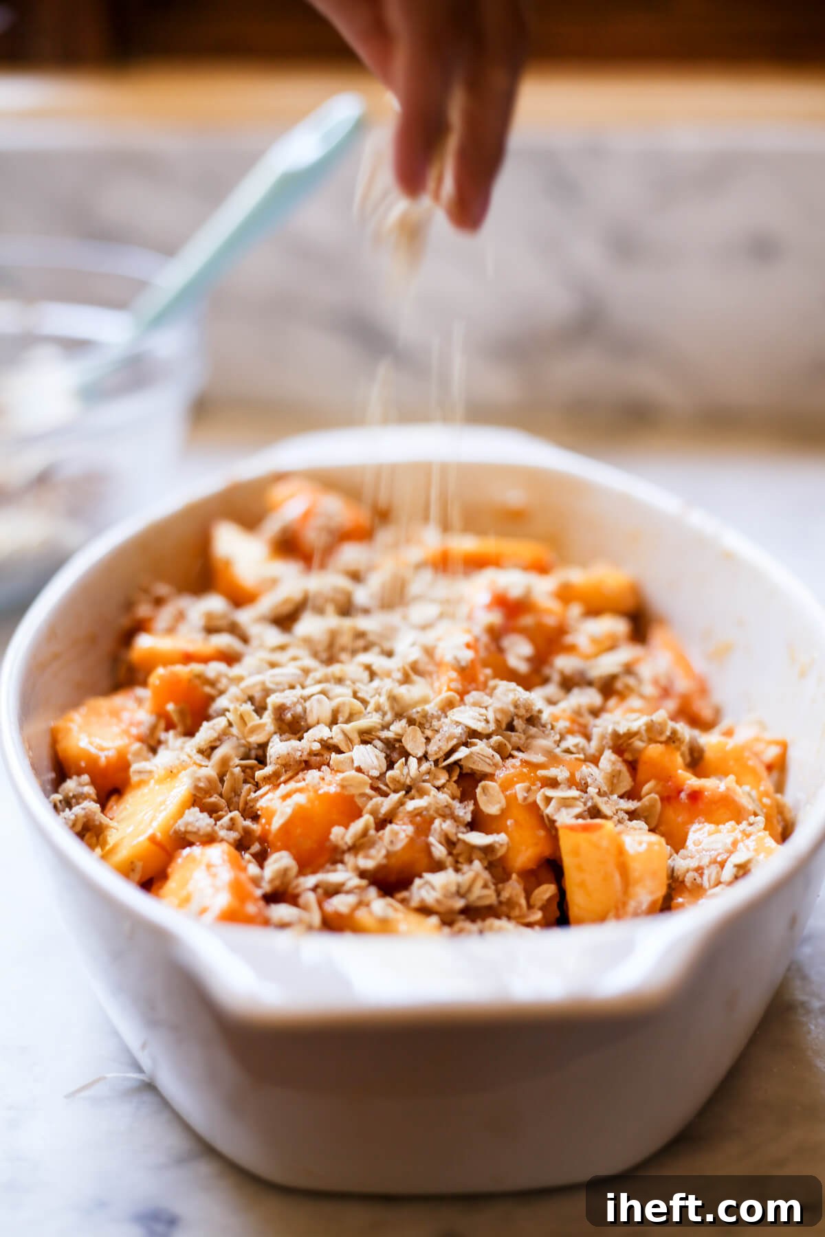 Oat crumble topping is sprinkled over peach crisp filling in a white oval ceramic baking dish.