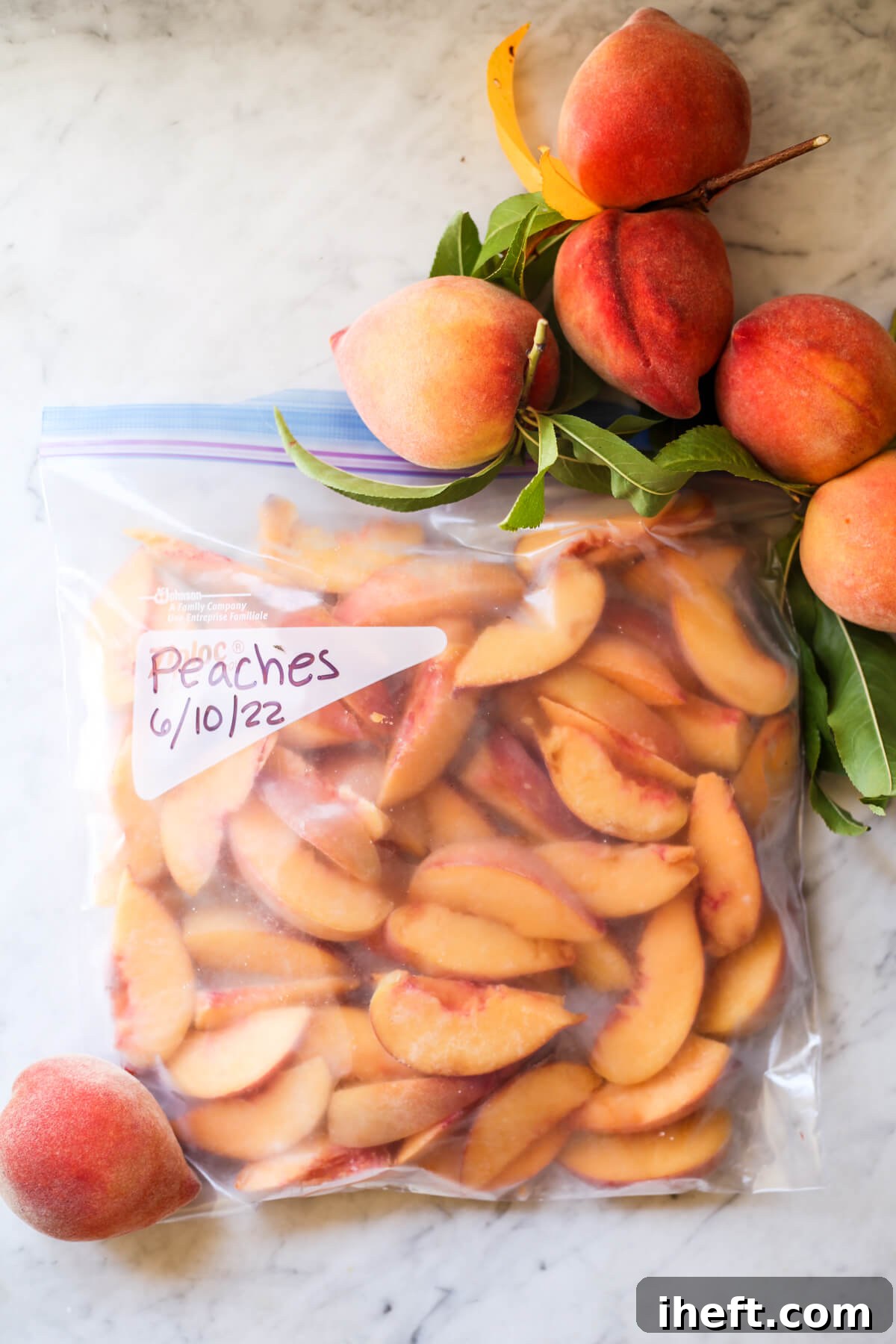 A freezer bag labeled "Peaches" is filled with frozen peach slices. The frozen peaches sit on a marble countertop with fresh whole homegrown peaches on the side. 