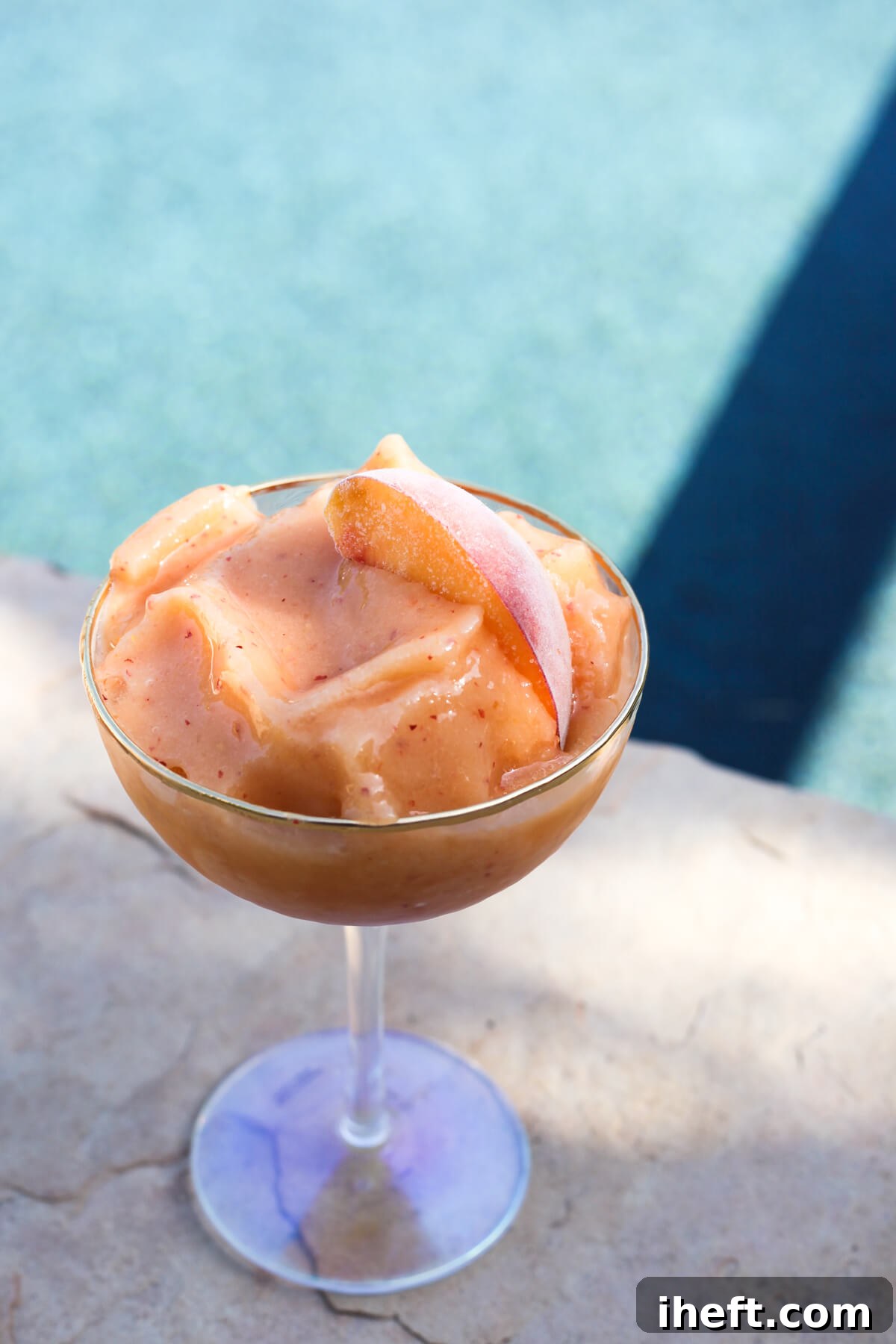 A coupe glass filled with a frozen peach smoothie in front of a pool. 