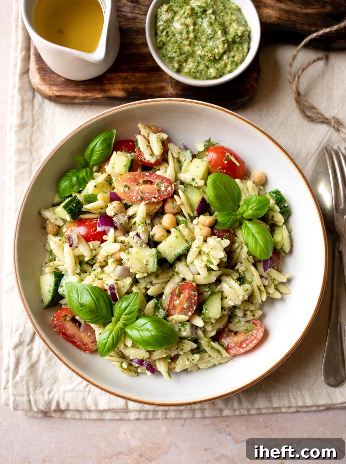 A beautifully presented bowl of pesto orzo salad, brimming with chickpeas, halved cherry tomatoes, fresh cucumber slices, and garnished with fragrant basil leaves. This is the epitome of a refreshing summer meal or side.