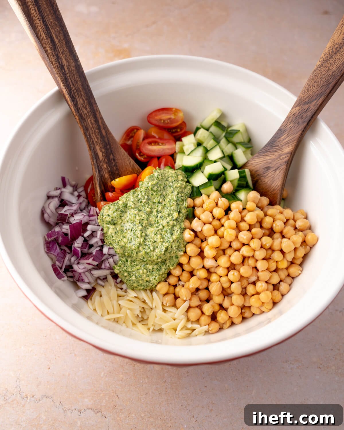 Ingredients for orzo salad are about to be mixed together with wooden spoons in a white bowl.