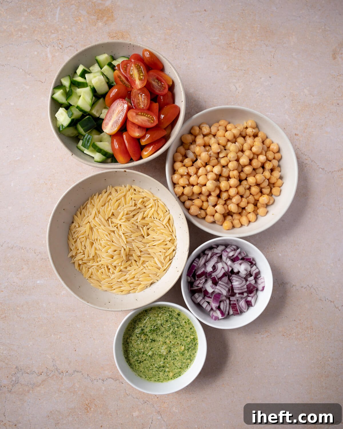 A curated display of fresh ingredients essential for crafting a delicious pesto orzo salad. The bowls, arranged clockwise, feature vibrant cucumber and tomatoes, protein-rich chickpeas, finely diced red onion, a generous serving of vibrant pesto, and dry orzo pasta ready for cooking.