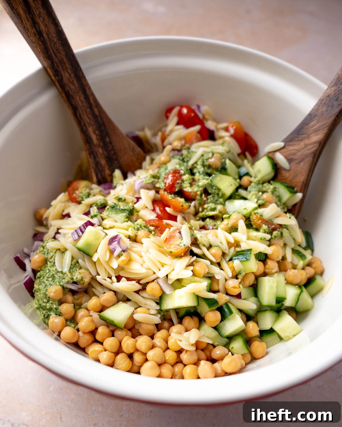 A large white salad bowl is filled with a colorful medley of cooked orzo, chickpeas, diced cucumber, halved cherry tomatoes, and finely chopped red onion. Two wooden spoons are poised, ready to toss and combine all the ingredients with a dollop of vibrant green pesto, illustrating the final steps of making a delicious pesto orzo salad.
