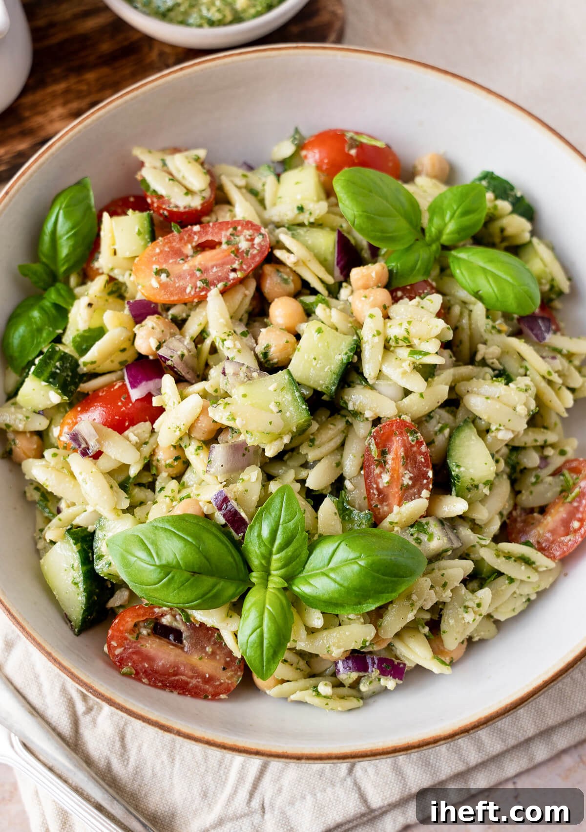 A close-up, inviting photo of a bowl filled with a delicious pesto orzo salad. The salad features plump chickpeas, vibrant cherry tomatoes, crisp cucumber, and fresh basil leaves, all glistening with a rich green pesto. It's a truly appetizing and visually appealing dish.