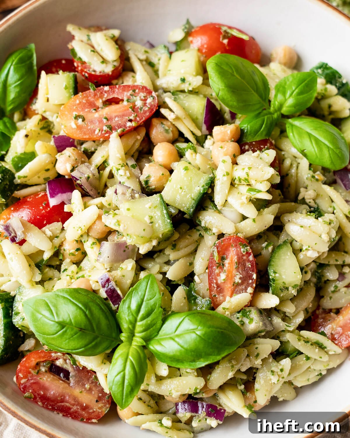 A tantalizing close-up shot of a summer orzo salad, prominently featuring the vibrant green pesto, juicy cherry tomatoes, crisp cucumber, plump chickpeas, and fragrant fresh basil leaves. This image highlights a delicious and visually appealing vegan pasta salad, perfect for warm weather.