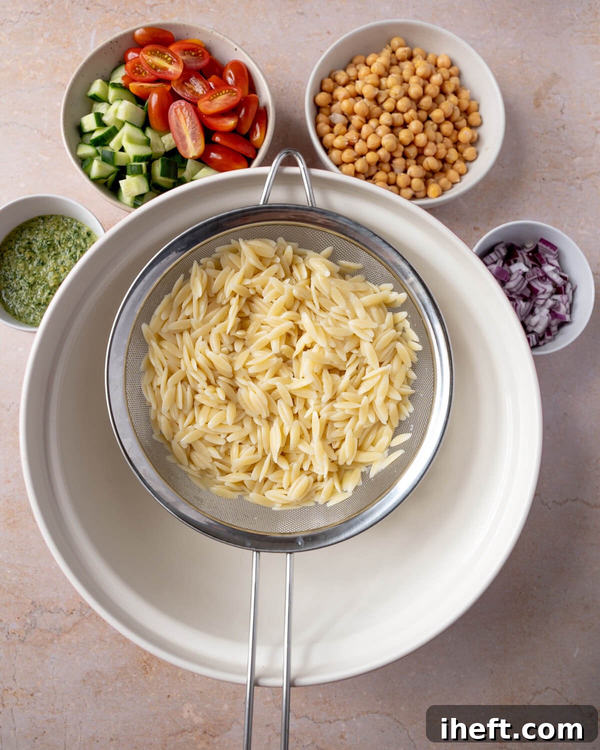 The ingredients for orzo salad around a white bowl. The orzo pasta salad ingredients include pesto, cucumber, tomatoes, chickpeas, and diced red onion.