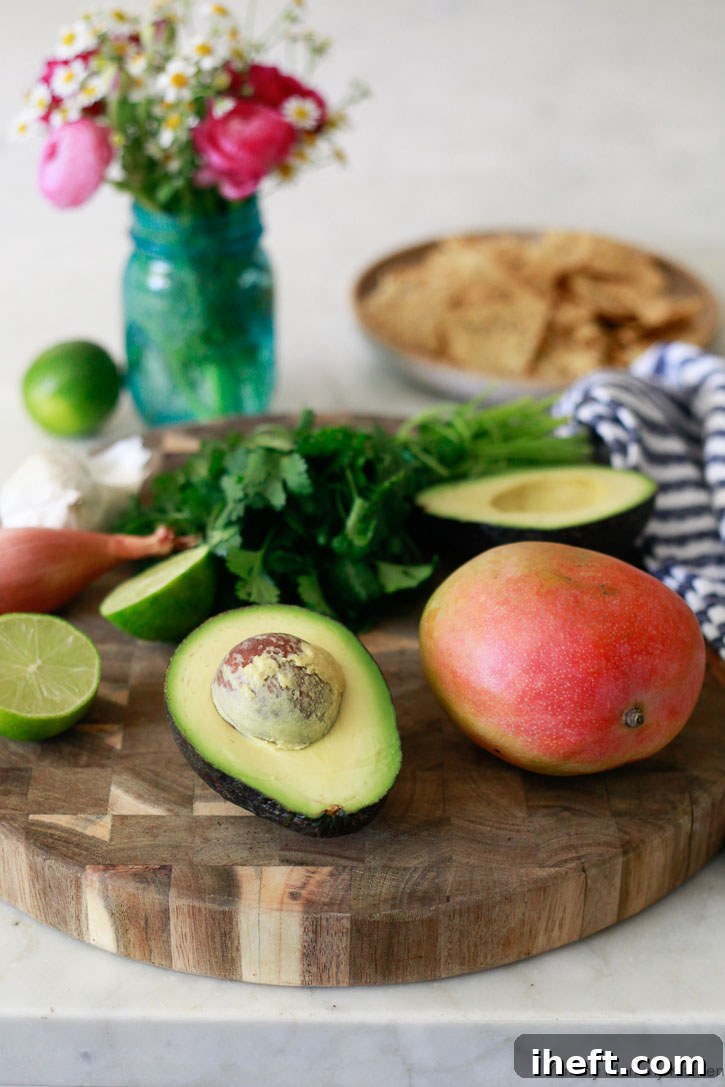 A close-up shot of the fresh ingredients for mango avocado salsa laid out on a clean surface, featuring a perfectly ripe mango, a creamy avocado, a small shallot, bright green limes, and a clove of garlic, ready for preparation.