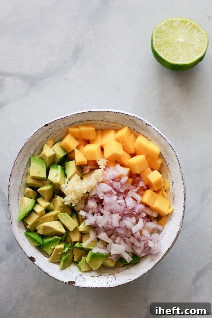 A clear glass bowl filled with the freshly diced ingredients for mango avocado salsa, including perfectly cubed avocado and mango, finely minced garlic, and delicate shallot, with vibrant lime wedges alongside, showcasing the beautiful preparation.