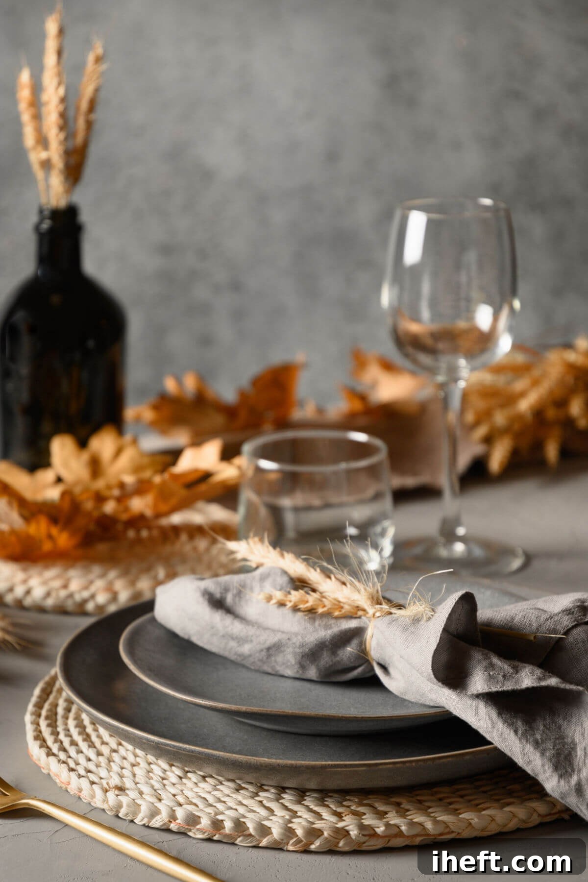 A chic grey and gold Thanksgiving table setting, featuring elegant grey stoneware plates, polished gold flatware, and a centerpiece crafted from dried wheat and delicate autumn leaves, creating a sophisticated modern fall aesthetic.