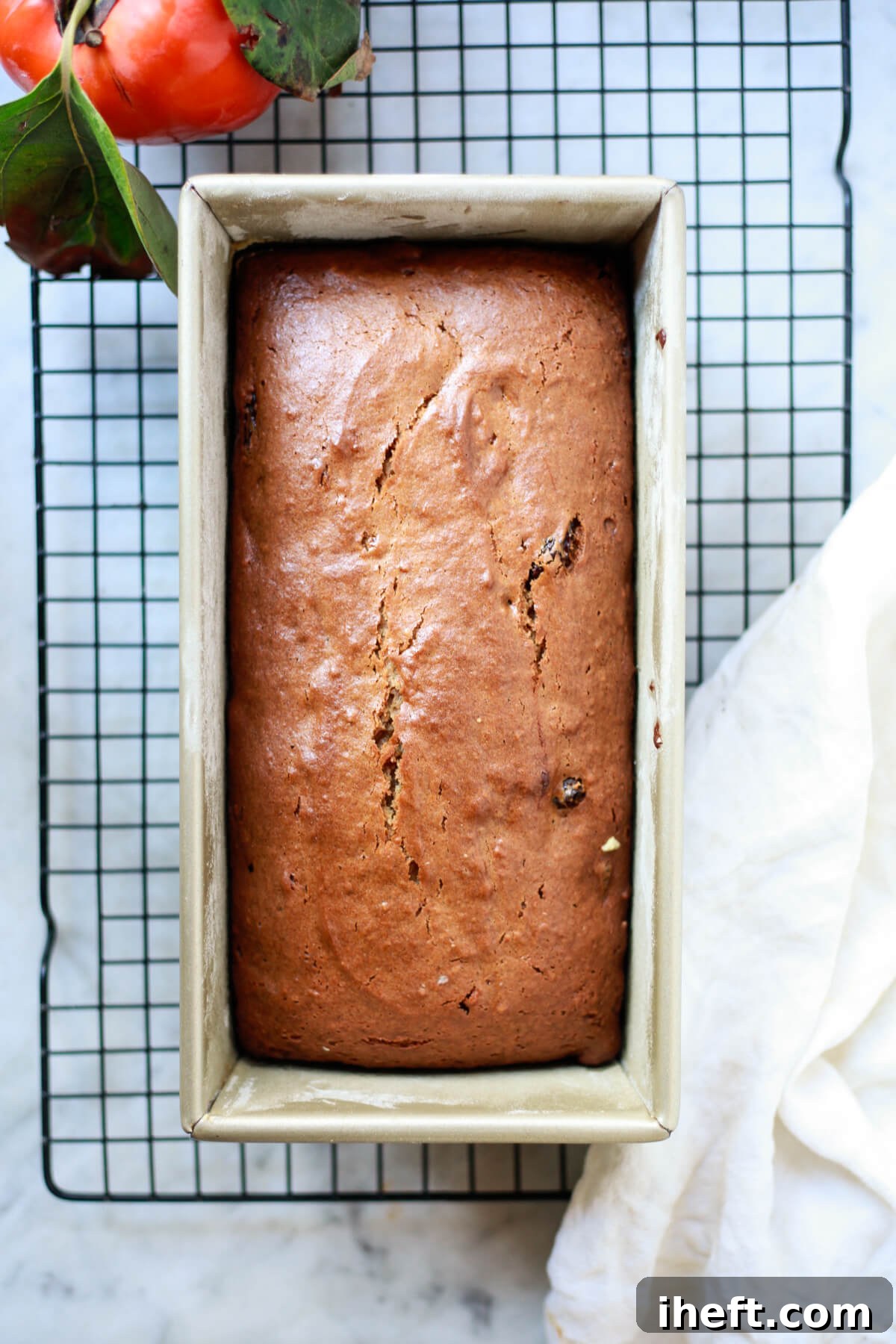 An overhead photo of golden-brown persimmon bread in a gold loaf pan on a wire cooling rack, ready to be sliced and enjoyed.