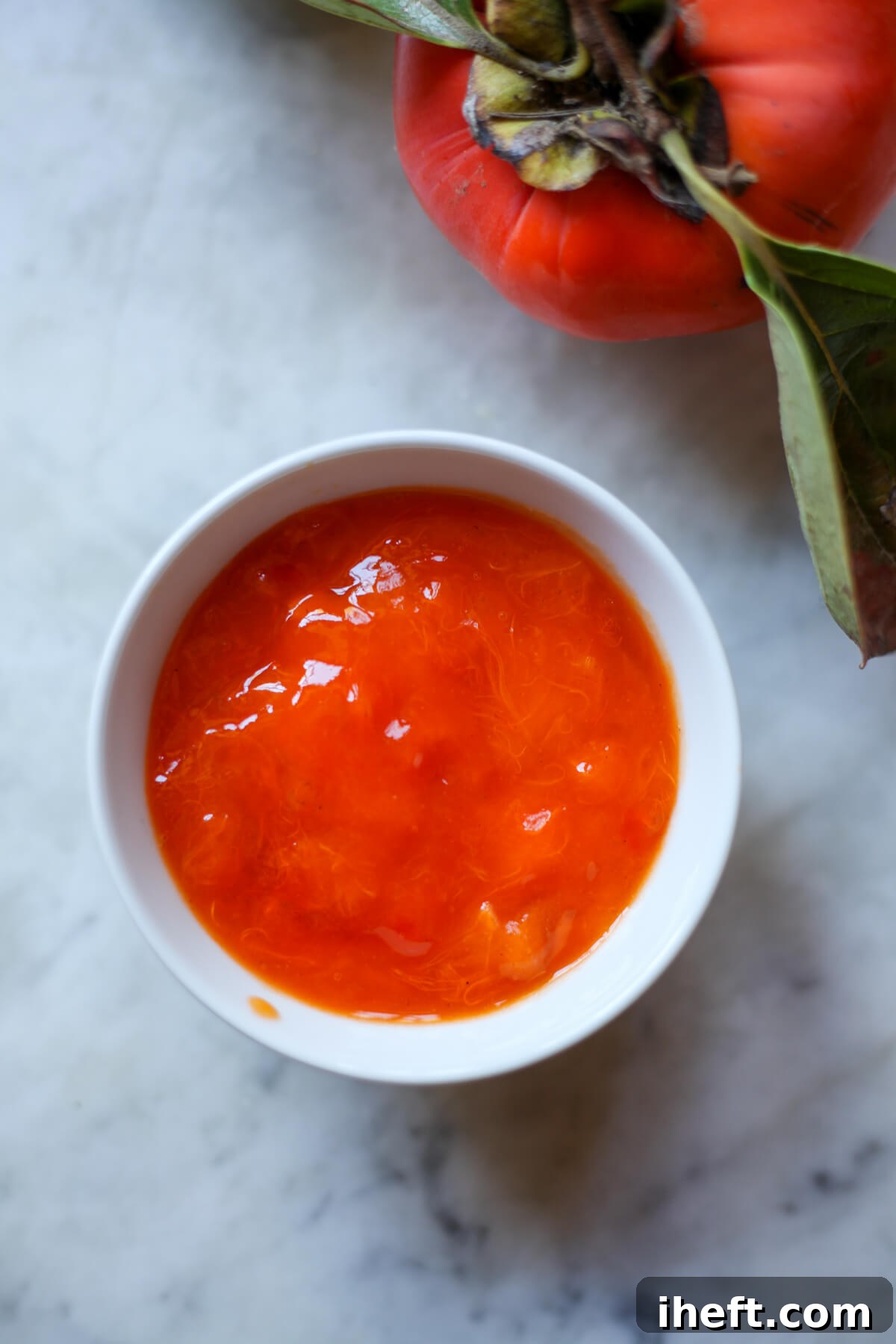 A small white bowl brimming with smooth, golden-orange Hachiya persimmon pulp or puree, resting on a cool marble countertop.