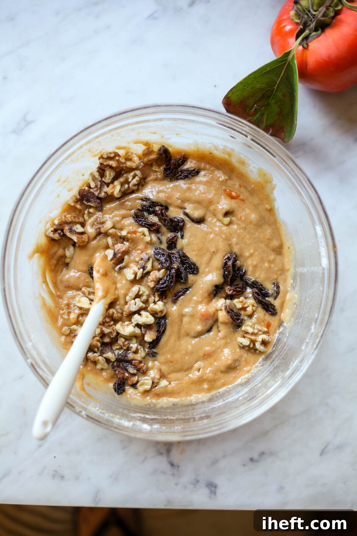Chopped walnuts and plump raisins are gently stirred into a rich, golden persimmon bread batter within a clear glass bowl, resting on a marble countertop.