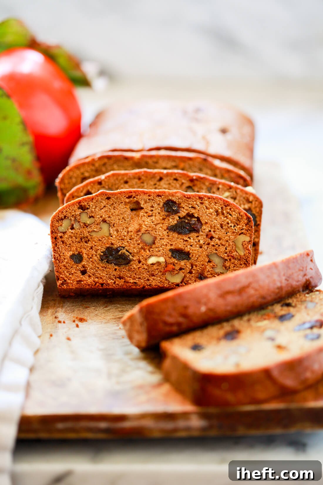 Slices of freshly baked persimmon bread, studded with raisins and walnuts, neatly arranged on a rustic wooden cutting board. A whole persimmon rests in the background on a white countertop.