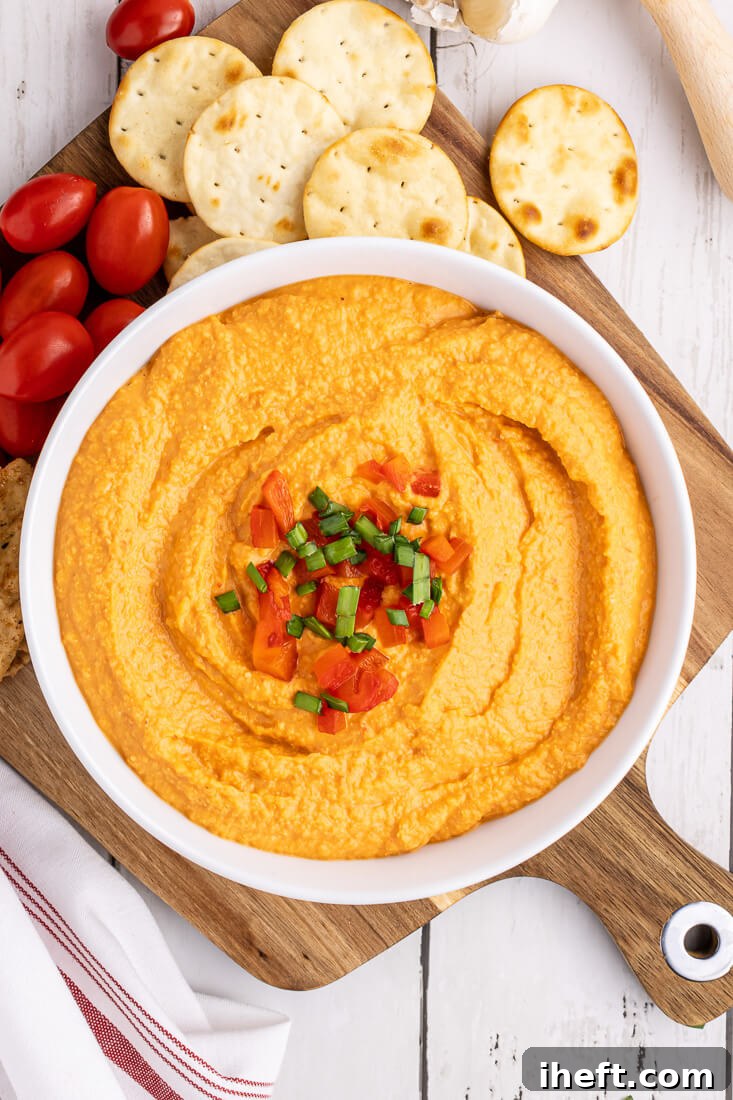 A white bowl filled with homemade roasted red pepper hummus on a wooden board served with crackers and tomatoes.