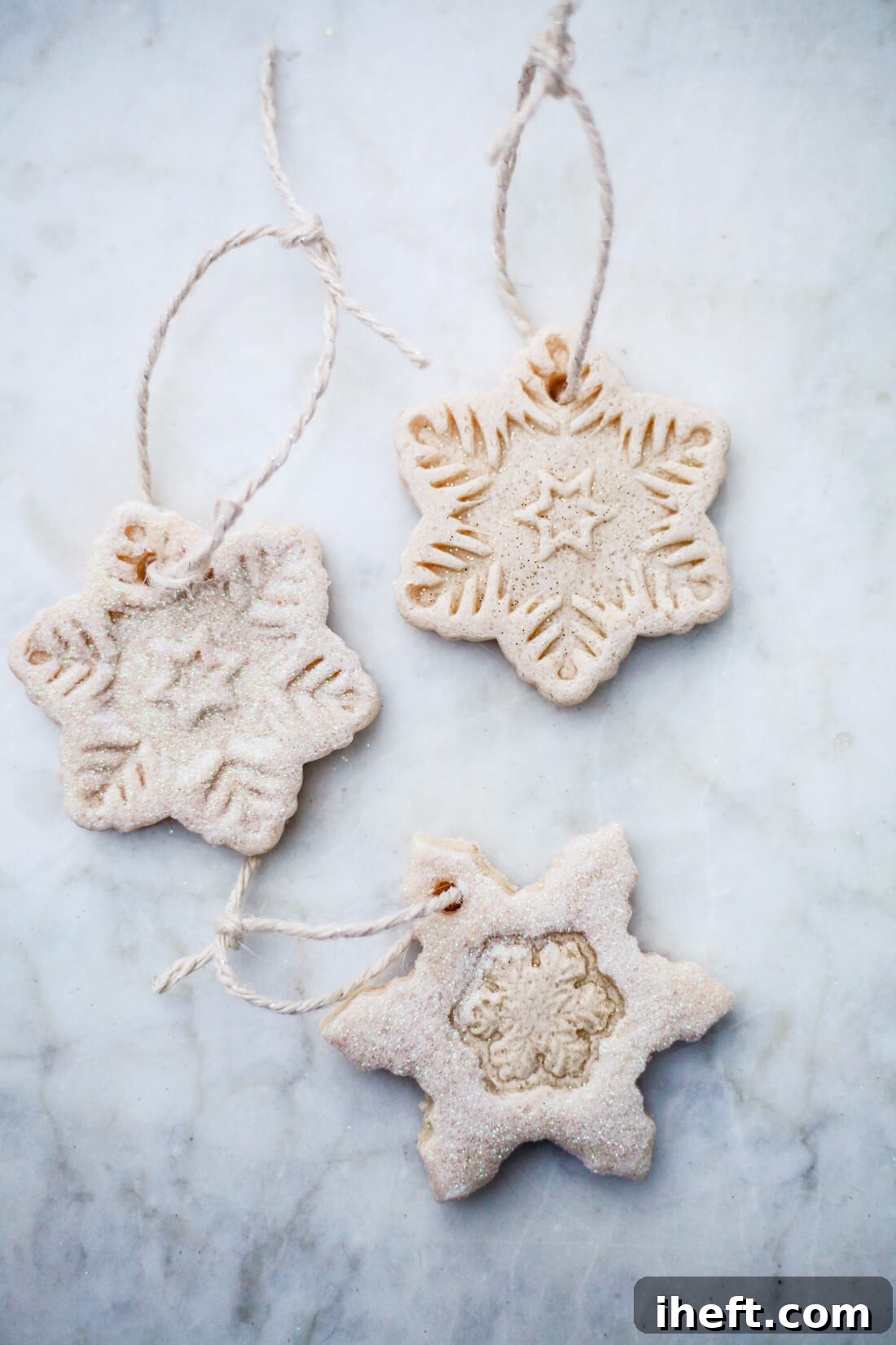 Three snowflake salt dough ornaments with twine and white glitter on a marble counter. 