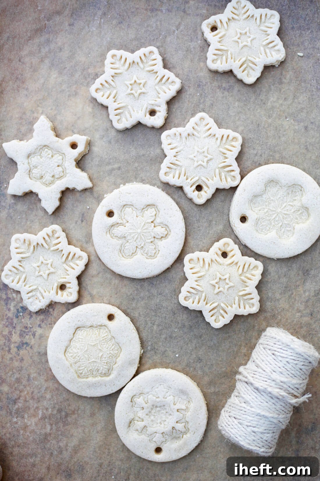 10 homemade salt dough ornaments on a baking sheet with a roll of twiine in the lower corner. These ornaments are white and in snowflake shapes. 