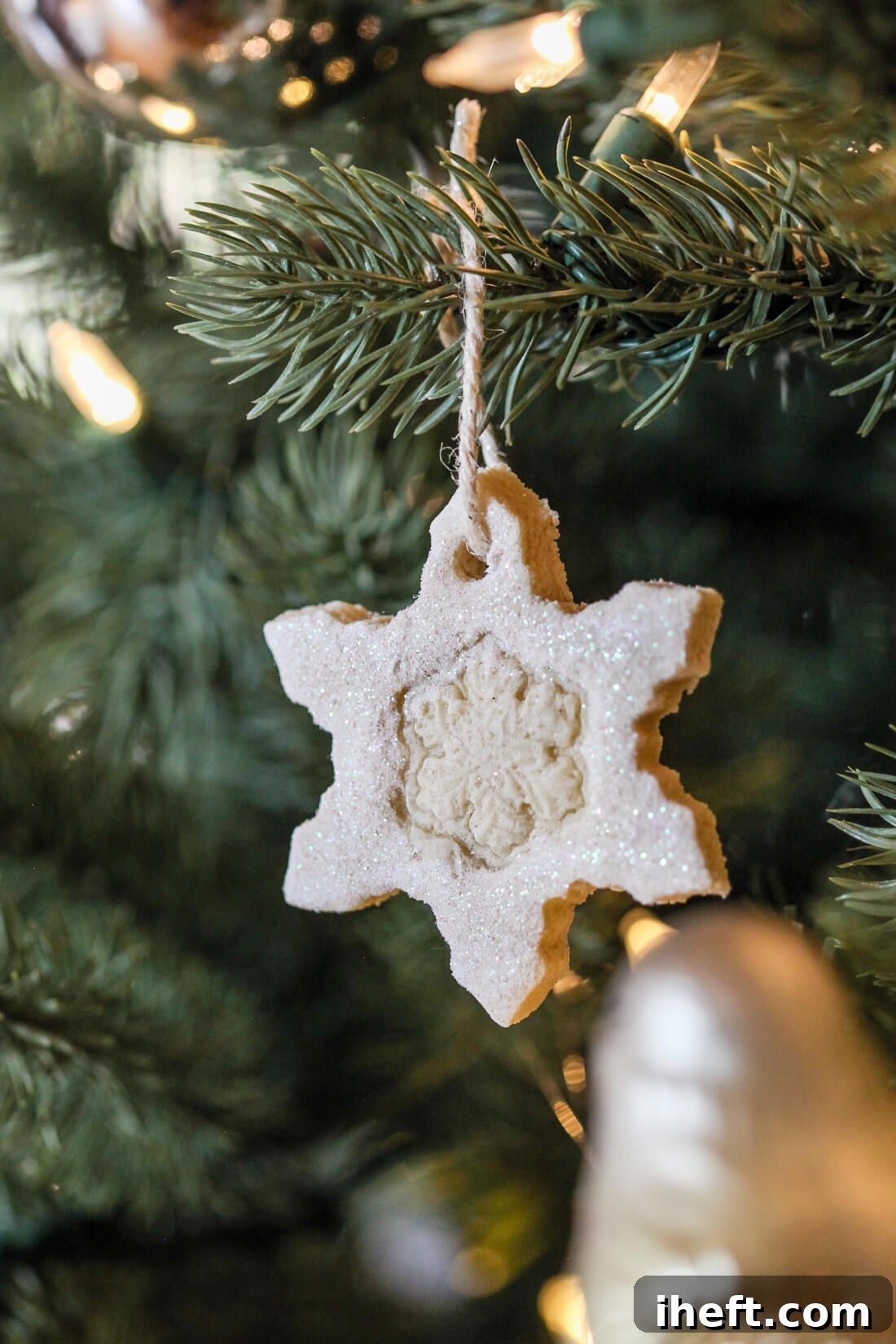 A white glitter coated salt dough snowflake ornament hangs from a Christmas tree branch by a pice of twine. 