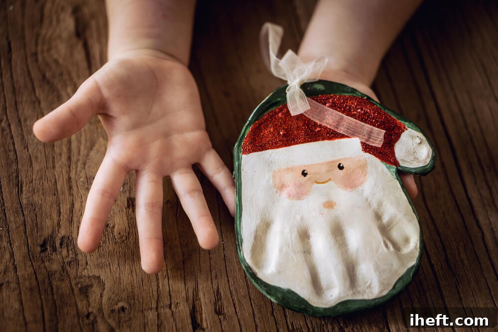 A child's hands hold a homemade salt dough ornament made with a handprint that has been painted to look like Santa with a red glitter Santa hat. 