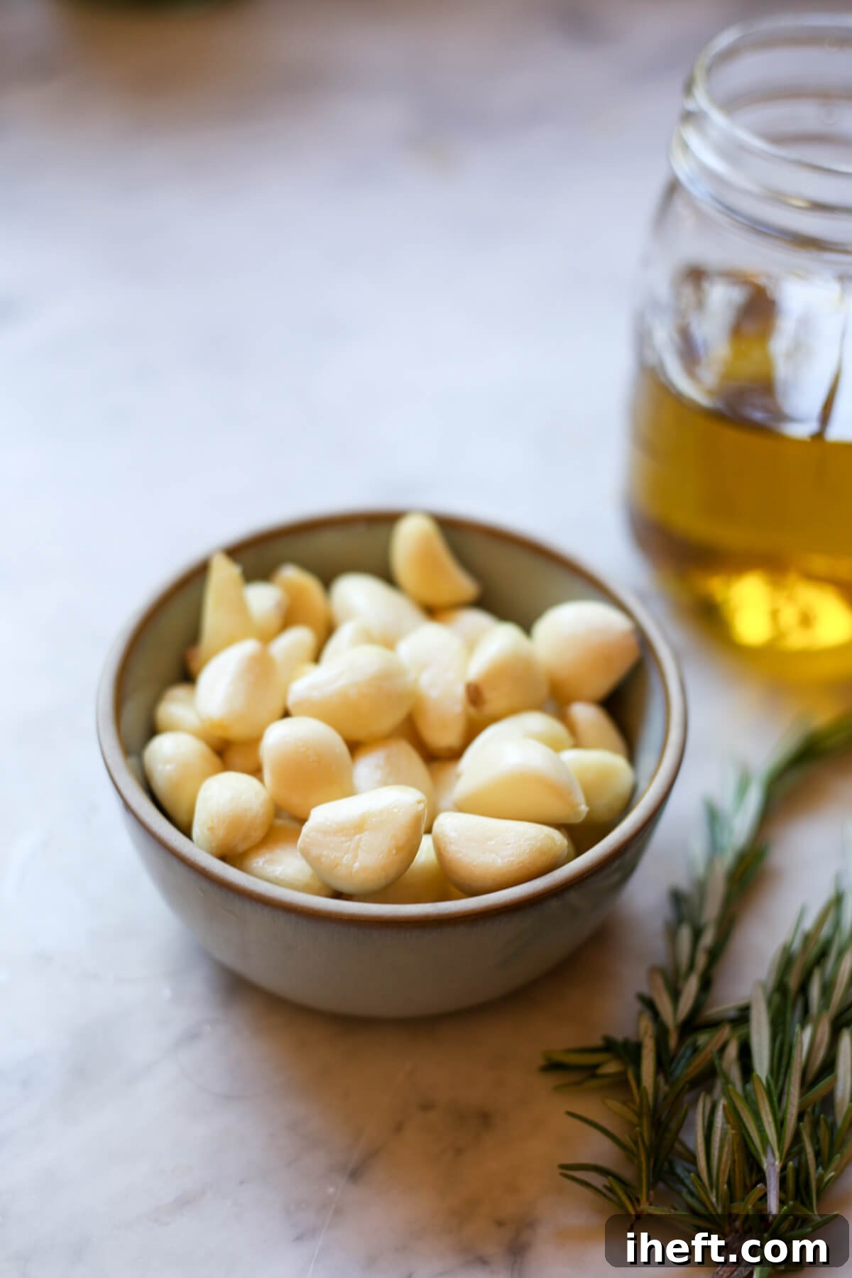 A small grey ceramic bowl filled with peeled garlic cloves on a marble counter with olive oil in the background and rosemary in the foreground.
