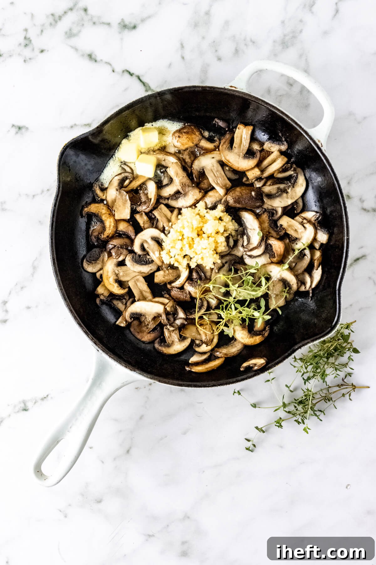 A skilled filled with sliced mushrooms, butter, minced garlic, and fresh thyme on a marble counter.