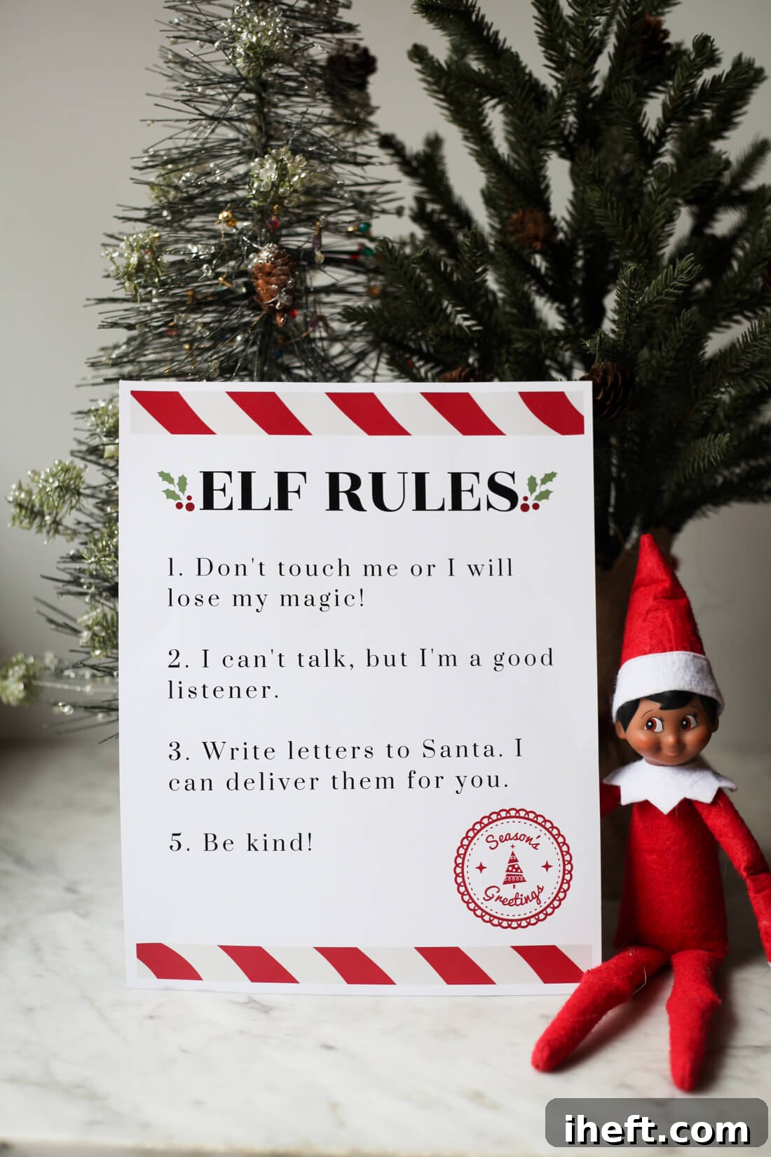An Elf on the Shelf sits next two two small Christmas trees and a printable that says "Elf Rules" with a list of the Elf on the Shelf Rules. 