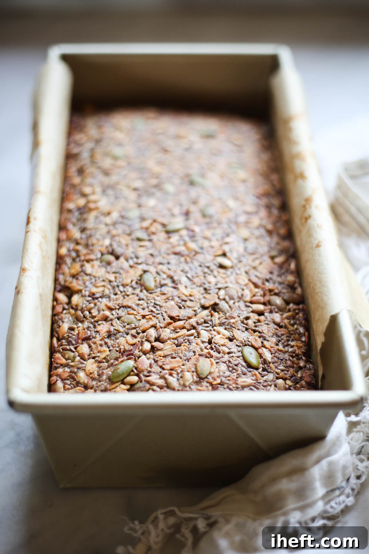 A freshly-baked loaf of super seed bread, golden brown, nestled in a gold loaf pan lined with parchment paper on a pristine white countertop. 