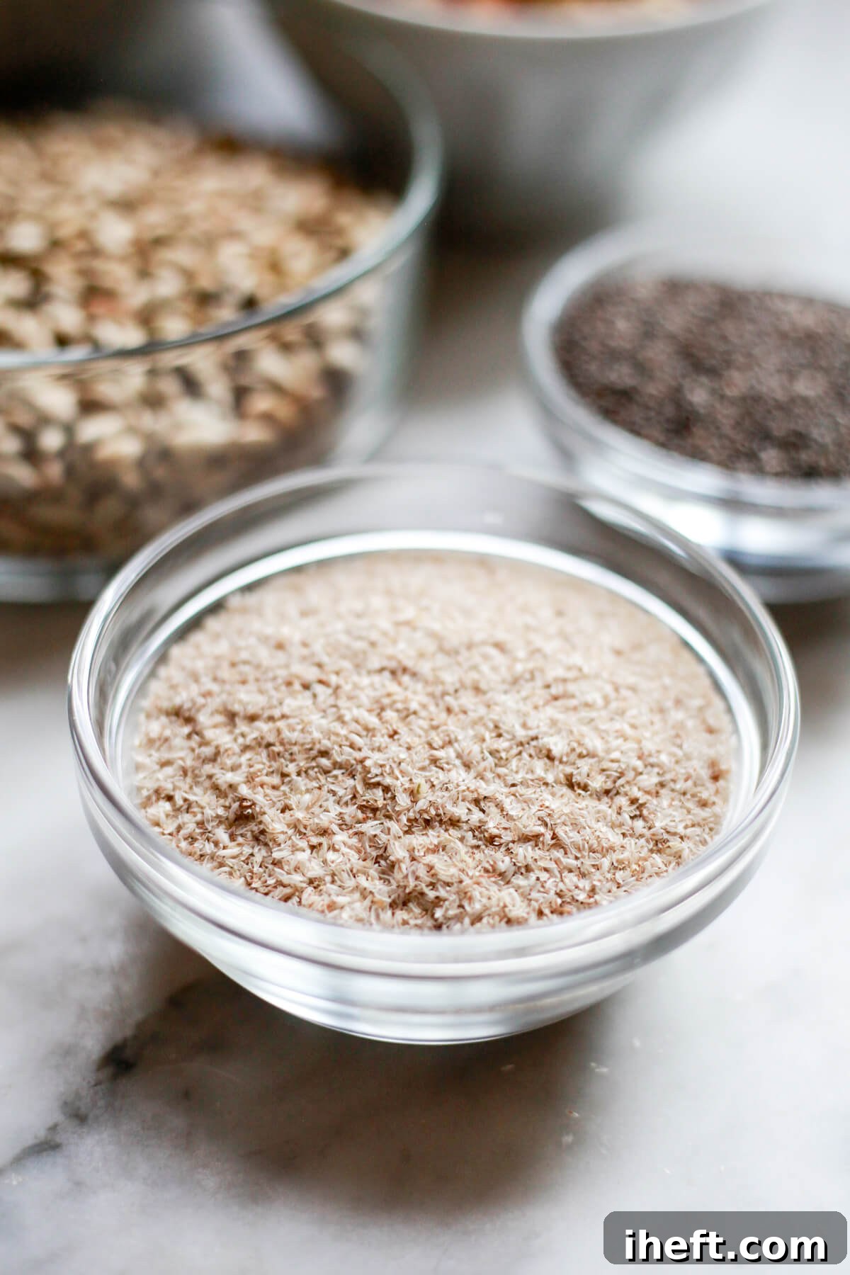 A small bowl brimming with psyllium husks rests on a sleek marble countertop, with other seeds softly blurred in the background, suggesting a holistic approach to nutrition. 