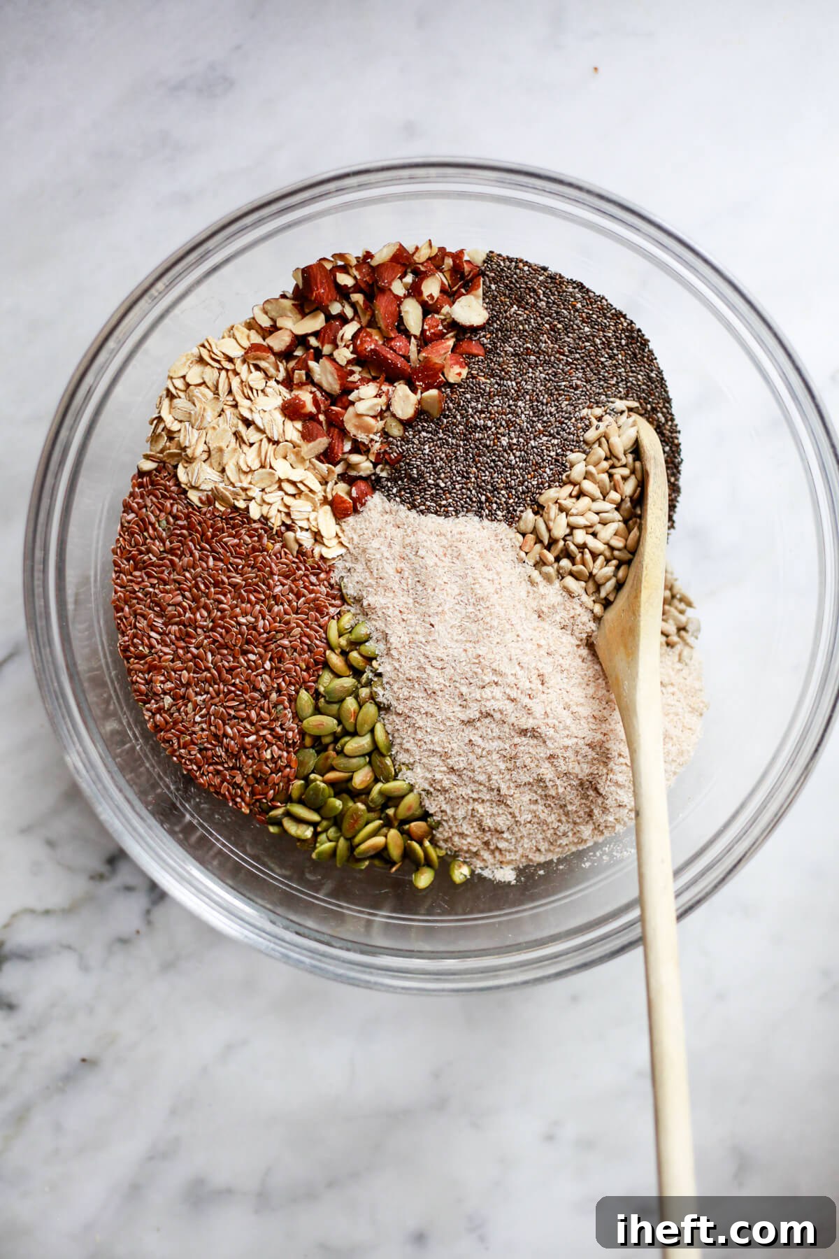 An overhead shot captures a large glass bowl, a harmonious blend of ingredients for seed bread, also known as adventure bread. The mix includes slivered almonds, tiny chia seeds, broad sunflower seeds, fluffy psyllium husks, dark pepitas, golden flax seeds, and rolled oats, all waiting to be transformed into a wholesome loaf. 