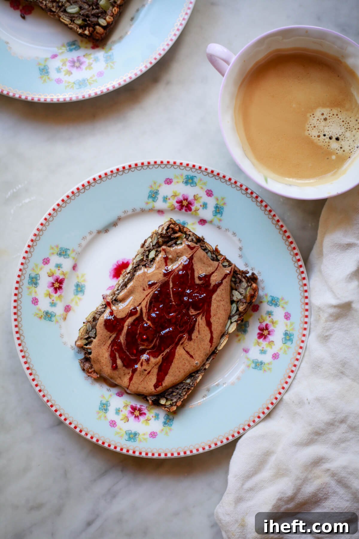 A charming scene featuring a slice of golden seed bread, generously topped with creamy peanut butter and sweet berry jam, resting on a floral plate. Beside it, a comforting cup of coffee completes this healthy, gluten-free, and vegan breakfast made with wholesome seed bread. 