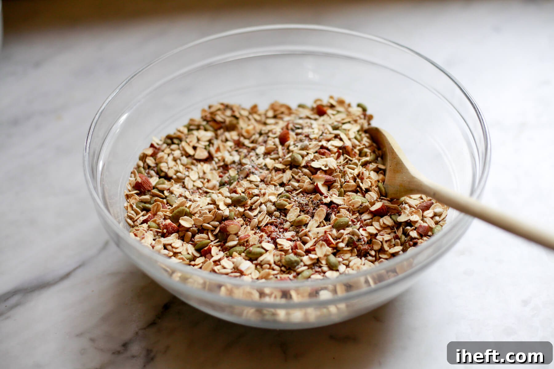 Seeds, nuts, and oats are stirred together in glass bowl with a wooden spoon.