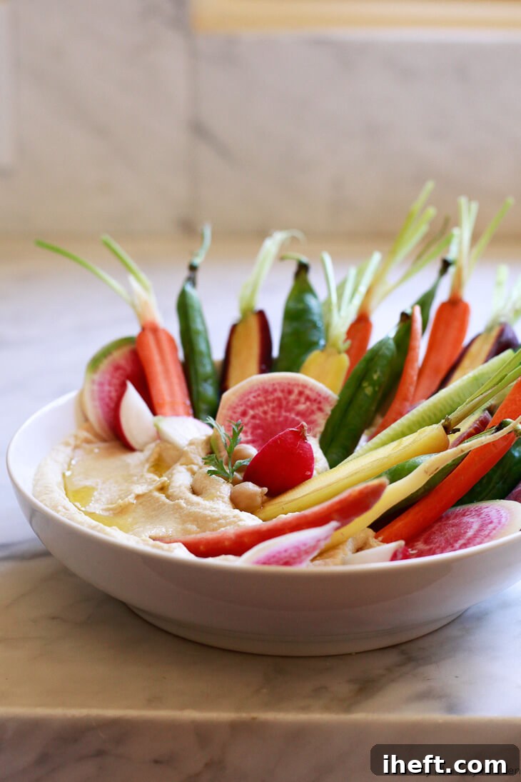 A beautifully garnished bowl of classic homemade hummus, served with an assortment of colorful fresh vegetables on a pristine white counter, ready for dipping.