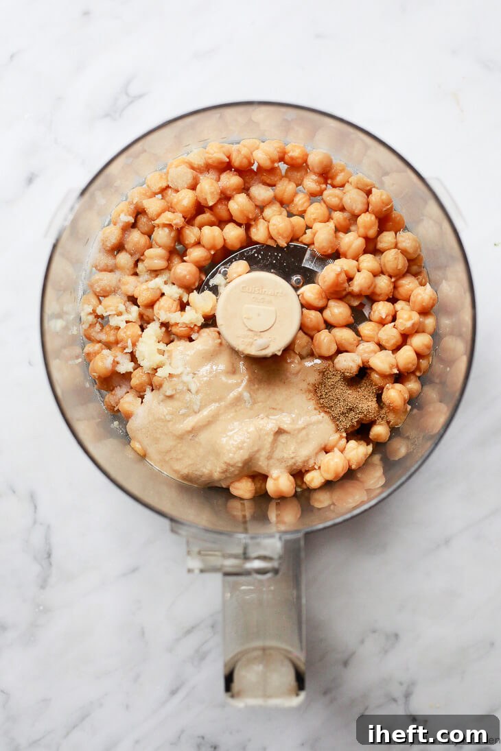 Close-up view of the key ingredients for homemade hummus, including chickpeas, creamy tahini, aromatic garlic, and earthy cumin, arranged in a food processor bowl before blending.